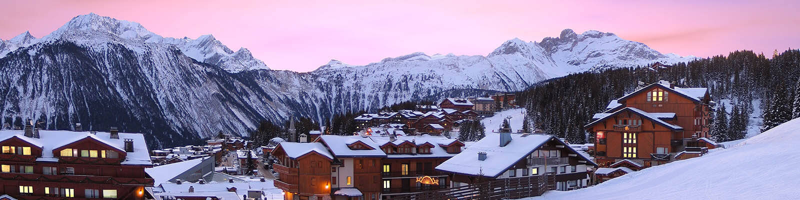 A scenic view of a snow-covered mountain village at sunset. Wooden chalets are scattered throughout the landscape, with snow blanketing the roofs. Majestic mountains loom in the background under a pink and purple sky.