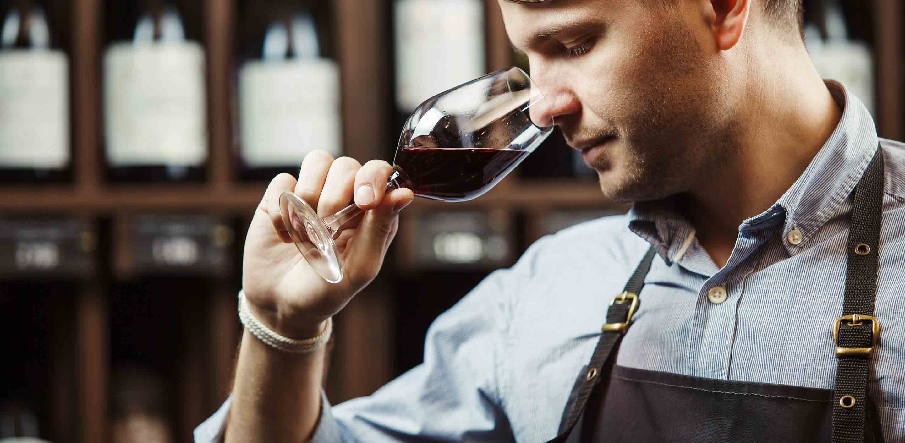 A man in a blue shirt and apron is holding a glass of red wine close to his nose, smelling it. He is standing in front of shelves stocked with wine bottles.