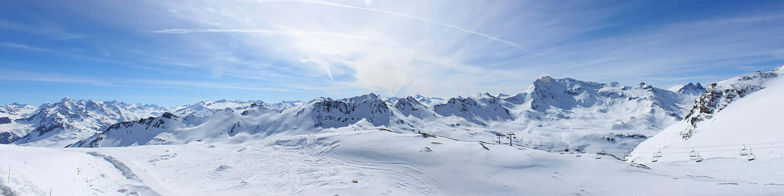 A breathtaking panoramic view of snow-covered mountains under a clear blue sky. Sunlight illuminates the peaks, casting subtle shadows. The landscape is vast and serene, with traces of ski tracks visible in the snow.