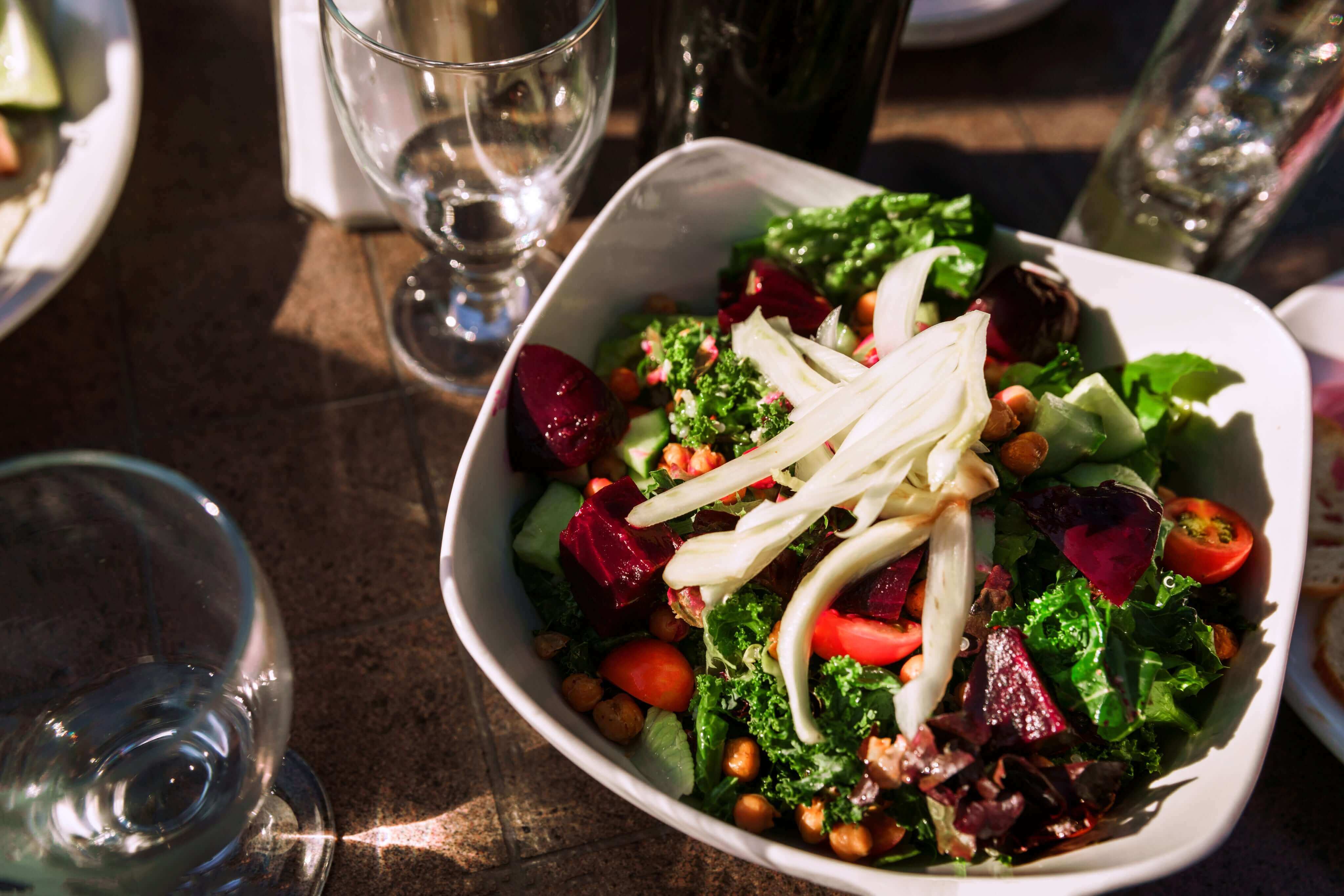 A vibrant salad in a white bowl featuring mixed greens, cherry tomatoes, beets, avocado, sliced fennel, and roasted chickpeas. The setting includes glasses and bottles on a sunlit wooden table.
