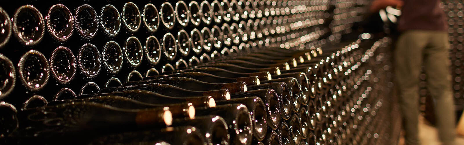 Rows of dusty, stacked wine bottles in a dimly lit cellar. The bottles are stored horizontally, forming a long, continuous line. The blurred figure of a person is visible in the background.