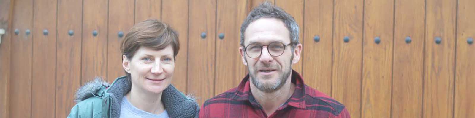 A woman and a man standing side by side in front of a wooden background. The woman wears a green jacket, and the man wears glasses and a red plaid shirt.