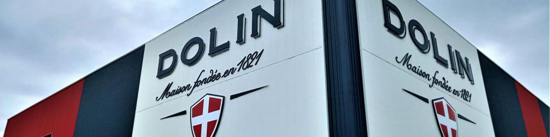 Corner of a building with a red, black, and gray facade. The sign reads DOLIN with a crest and Maison fondée en 1815. Sky is overcast.