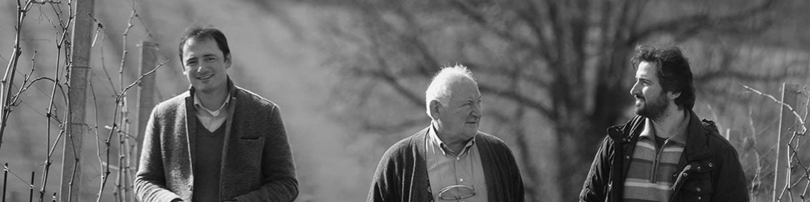 Three men walking and smiling outdoors in a vineyard. The background features bare vines and a large tree. The image is in black and white.