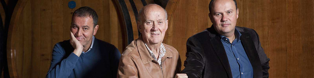 Three men are sitting and smiling in front of large wooden barrels. The man on the left rests his hand on his chin, the man in the center wears a brown jacket, and the man on the right wears a blue shirt and dark blazer.