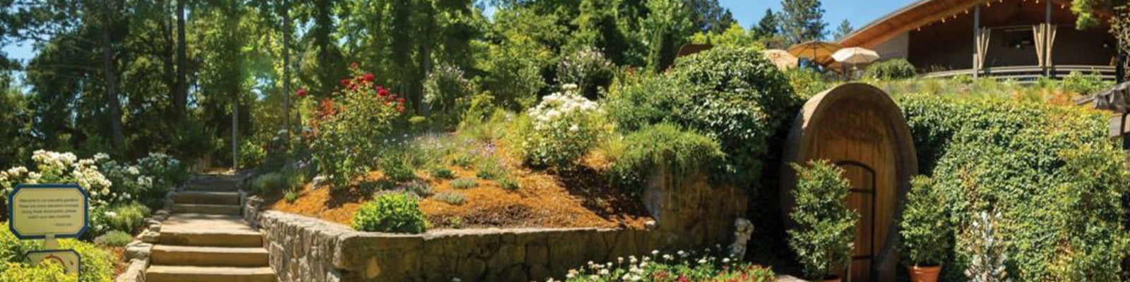 A picturesque garden with stone steps leading up to a large wooden door set in a hill. Colorful flowers and lush greenery surround the walkway, and a wooden house with umbrellas is in the background.