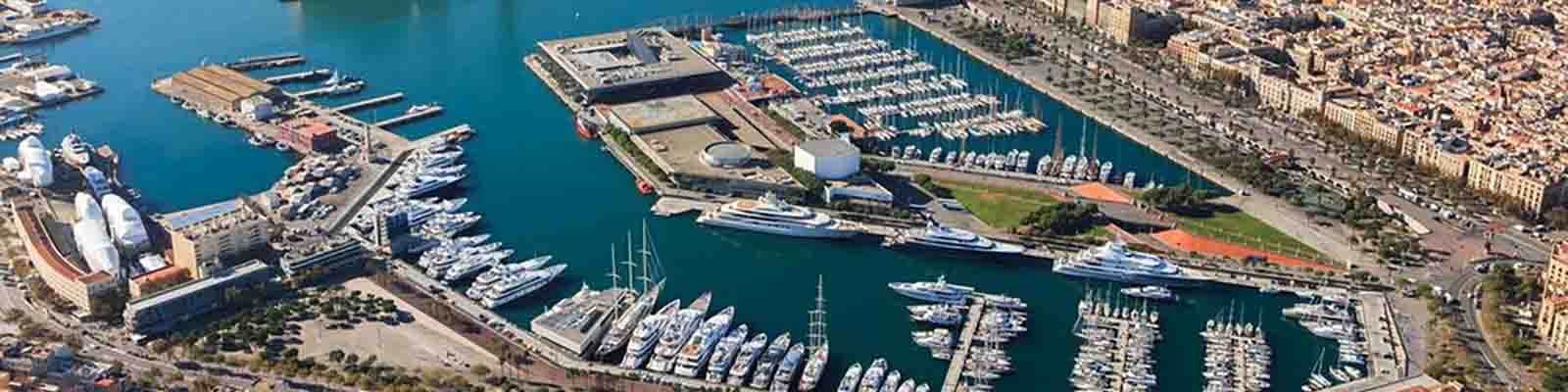 Aerial view of a bustling marina with numerous boats and yachts docked in a large harbor. The surrounding area features buildings, roads, and lush green spaces, indicating a lively coastal city.