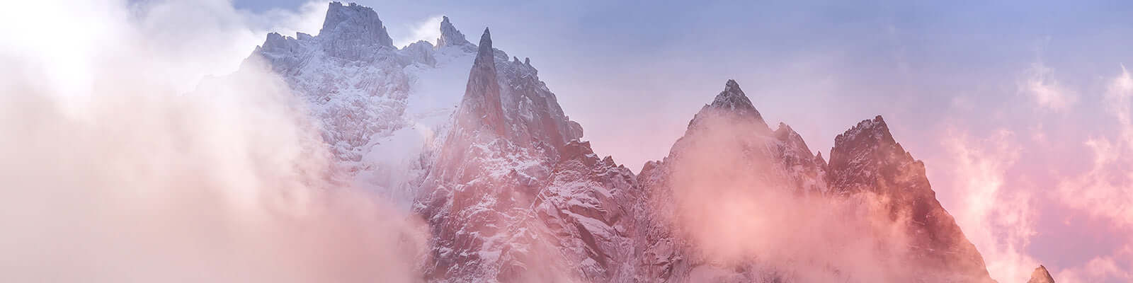 A panoramic view of snow-covered mountain peaks shrouded in pink and orange mist, under a soft sky transitioning from blue to pastel hues at sunrise or sunset.
