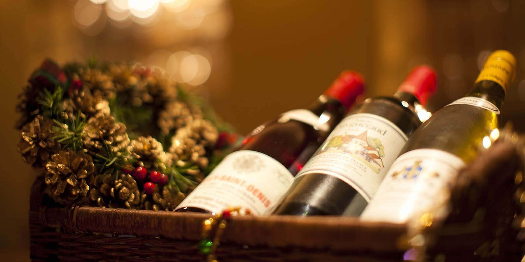 A festive wicker basket contains three bottles of wine, adorned with a holiday wreath made of pine cones and red berries. The scene is warmly lit, creating a cozy and inviting atmosphere.