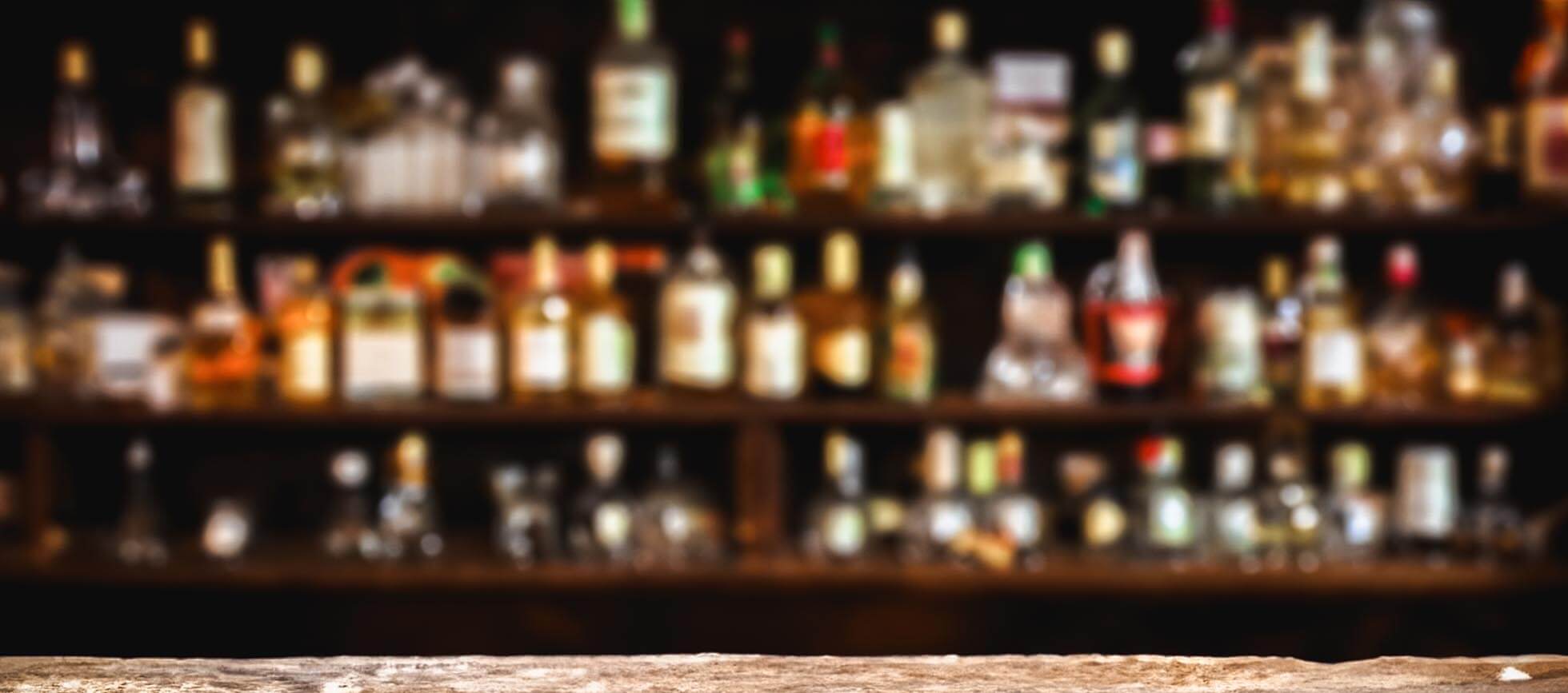 A blurred background showcases a variety of colorful liquor bottles displayed on wooden shelves, with a rustic wooden counter in the foreground.