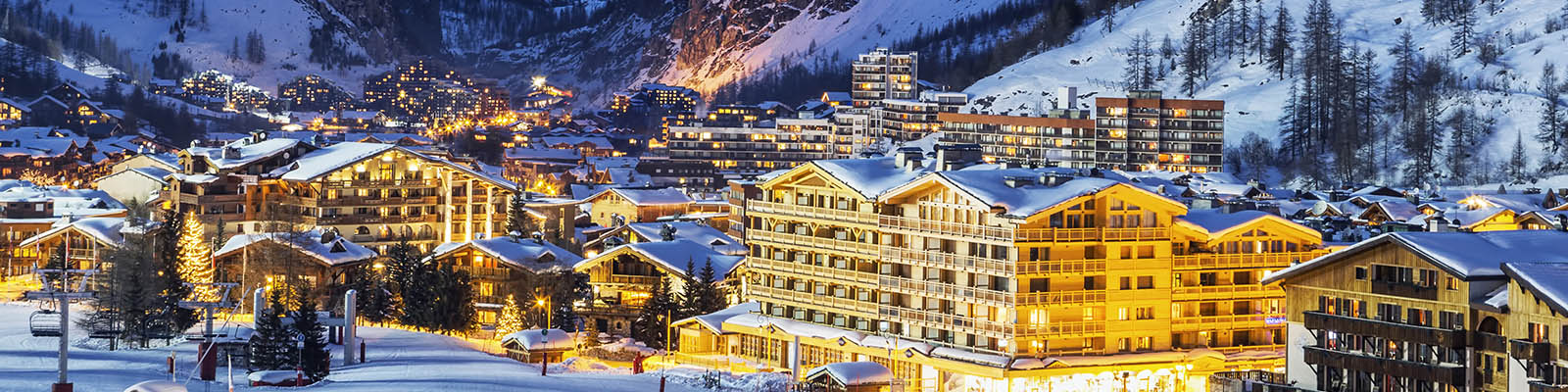 Night view of a snowy alpine village with warmly lit wooden buildings. Snow-covered roofs and trees are visible, surrounded by mountains in the background under a clear sky.