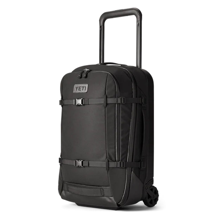 A Yeti Crossroads Luggage 22 in black, featuring a hard shell design with a retractable handle and sturdy wheels, is depicted. The suitcase includes front buckles and straps for secure closure and is shown positioned upright against a white background.