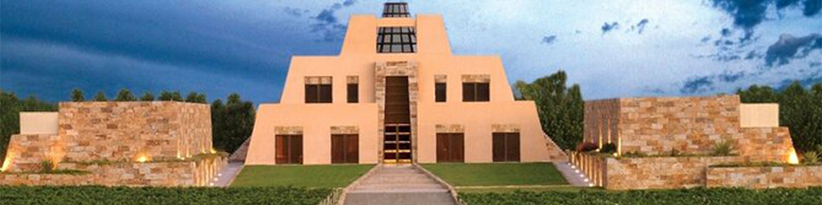 A modern building with a stepped pyramid shape, surrounded by greenery and flanked by stone walls under a cloudy sky.