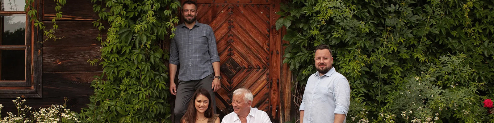 Four people posing in front of a wooden door surrounded by lush greenery and flowers. The group includes three men and one woman, all smiling.