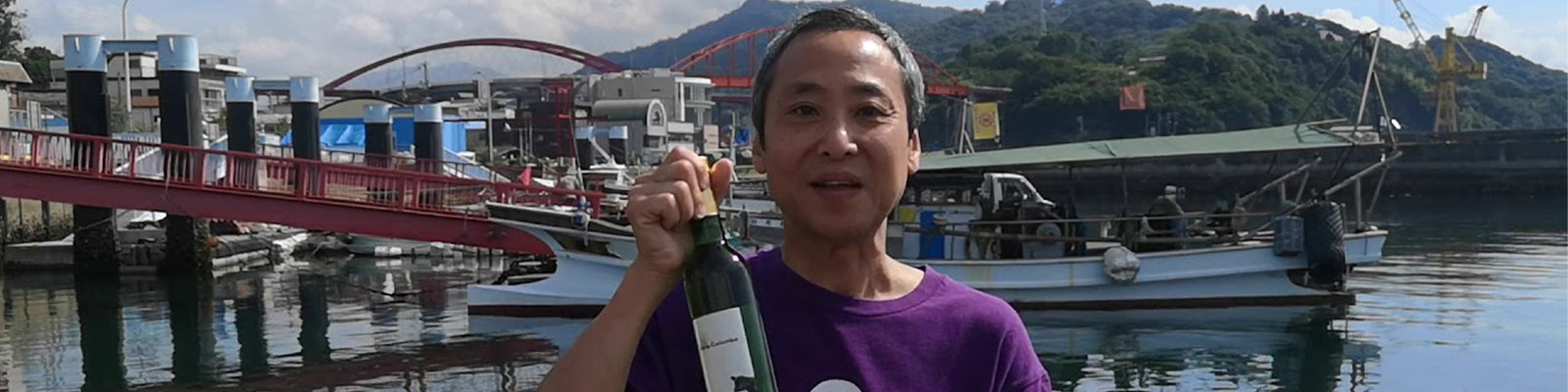 A person in a purple shirt holds a bottle while standing by a waterfront, with boats and a red bridge in the background. Hills and a partly cloudy sky are visible.