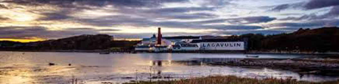 Ein malerischer Blick auf die Lagavulin-Brennerei vor einem dramatischen Himmel mit Wolken bei Sonnenuntergang. Das Gebäude liegt am Wasser, mit sichtbaren Spiegelungen auf der Wasseroberfläche, und ist von einer natürlichen Landschaft umgeben.