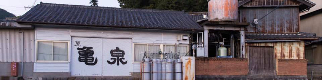 A traditional Japanese building with a tiled roof, featuring large kanji characters on a white facade. Industrial tanks and machinery are visible to the right, set against a backdrop of trees and mountain.