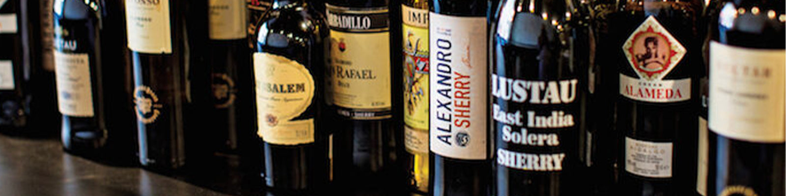 A row of various wine and sherry bottles with diverse labels displayed on a dark surface.