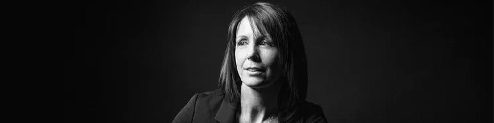 Black and white portrait of a woman with shoulder-length hair, wearing a blazer. She is looking slightly to the side against a dark background.