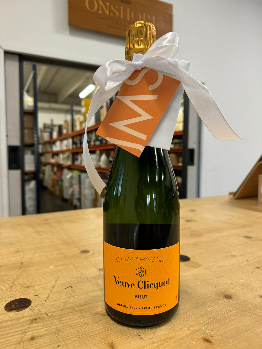 Eine Flasche Veuve Clicquot Brut Champagner mit einer weißen Schleife und einem orangefarbenen Etikett, die auf einem Holztisch steht. Im Hintergrund ist ein Lager mit Regalen und Kisten zu sehen.