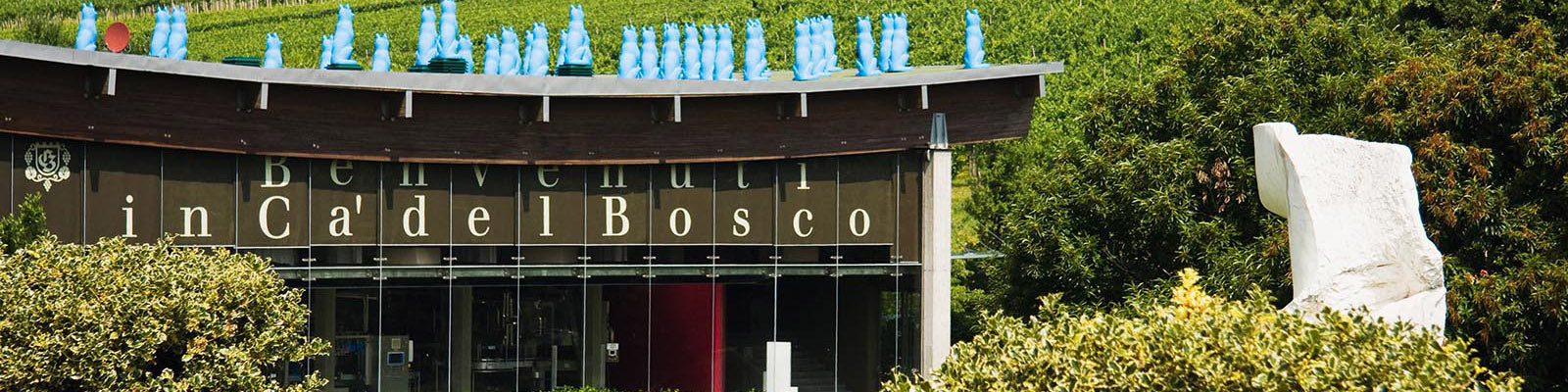 Modern building surrounded by greenery with Benvenuti in Ca del Bosco written on it. Blue sculptures adorn the roof, and a white sculpture is visible nearby.