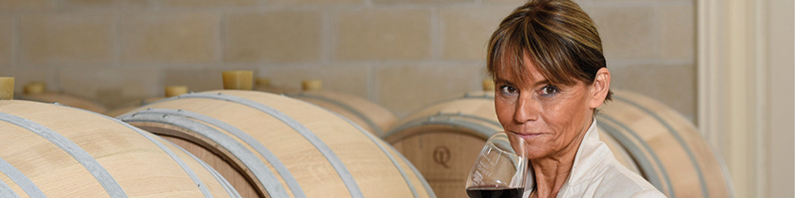 A person holding a glass of red wine stands in front of large wooden barrels in a winery.