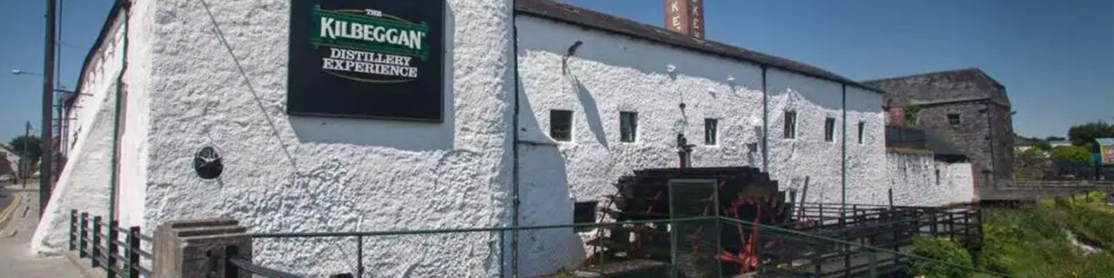 Exterior view of the Kilbeggan Distillery, featuring whitewashed stone walls, a sign reading Kilbeggan Distillery Experience, and an old waterwheel structure in the foreground under a blue sky.