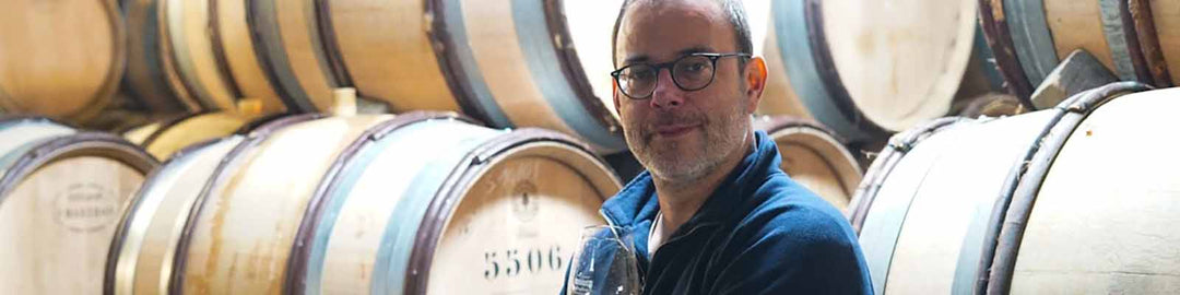 A person holding a wine glass stands in front of stacked wooden barrels in a winery.