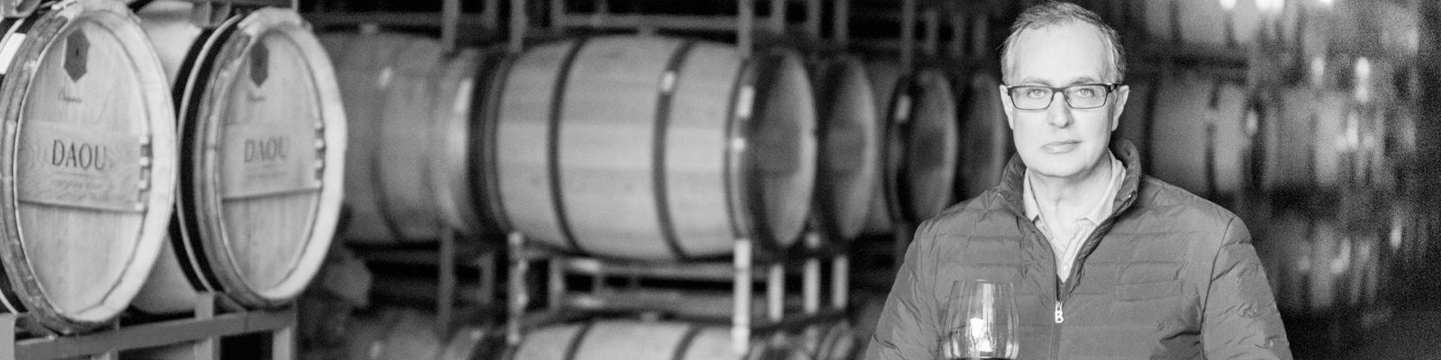 A person in a winery stands holding a glass of wine with wooden barrels stacked behind them. The setting is dimly lit, creating a classic and traditional atmosphere.