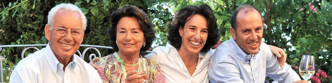 A group of four adults, two men and two women, sitting closely together outdoors, smiling and holding glasses of wine. Lush greenery is visible in the background.