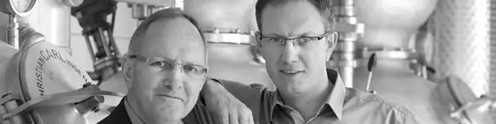 Two men wearing glasses stand side by side in front of industrial equipment. One has his hand on the others shoulder. The background includes large metal machinery. The image is in black and white.