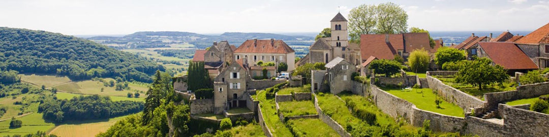 A picturesque view of a quaint village with stone houses and a church, surrounded by lush green hills and countryside under a clear blue sky.