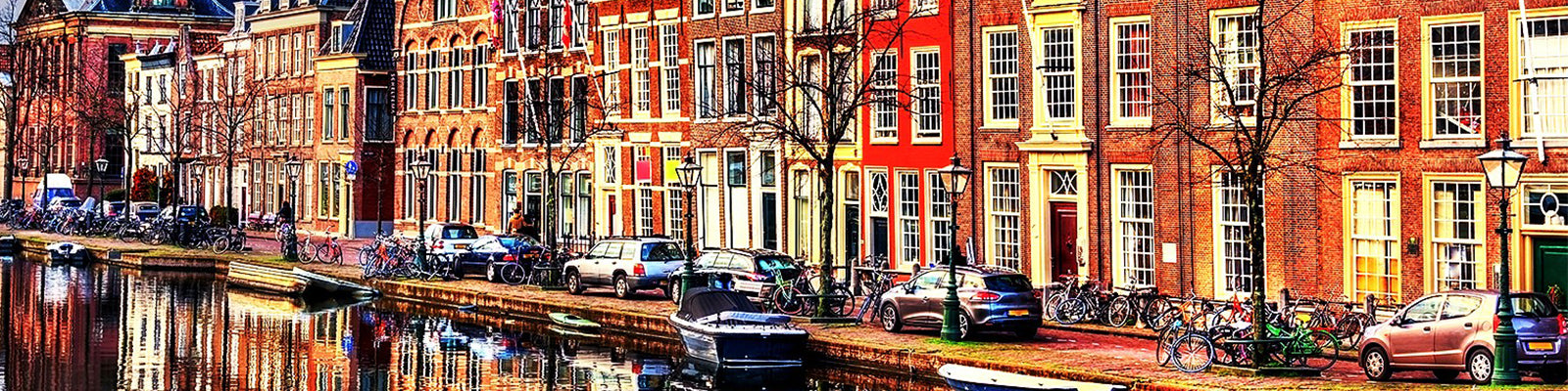 Colorful canal houses lined up along the water with bicycles and parked cars in front. A small boat is on the canal, and leafless trees stand in front of the buildings under a bright sky.