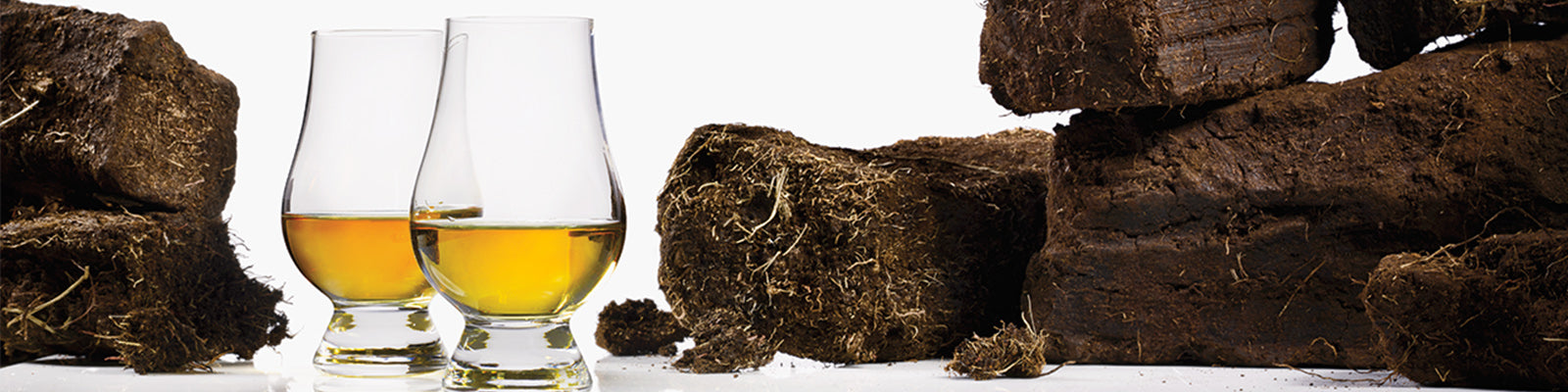 Two glasses of amber whiskey sit beside stacks of dark brown peat bricks on a white background.