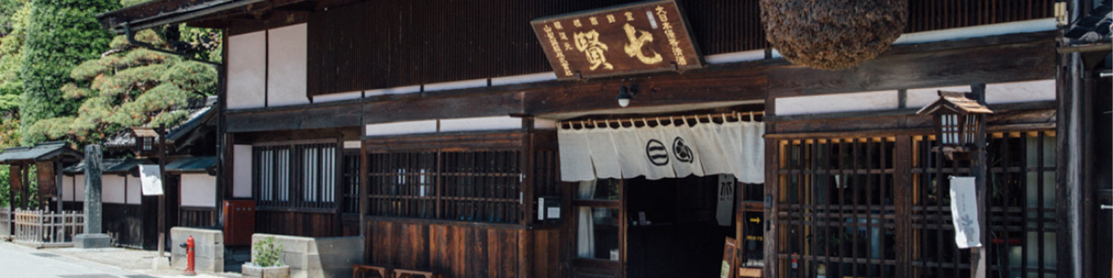 Traditional Japanese wooden building with sliding doors and a hanging sign. A white noren curtain with black symbols hangs at the entrance. Lush greenery is visible on the left side.