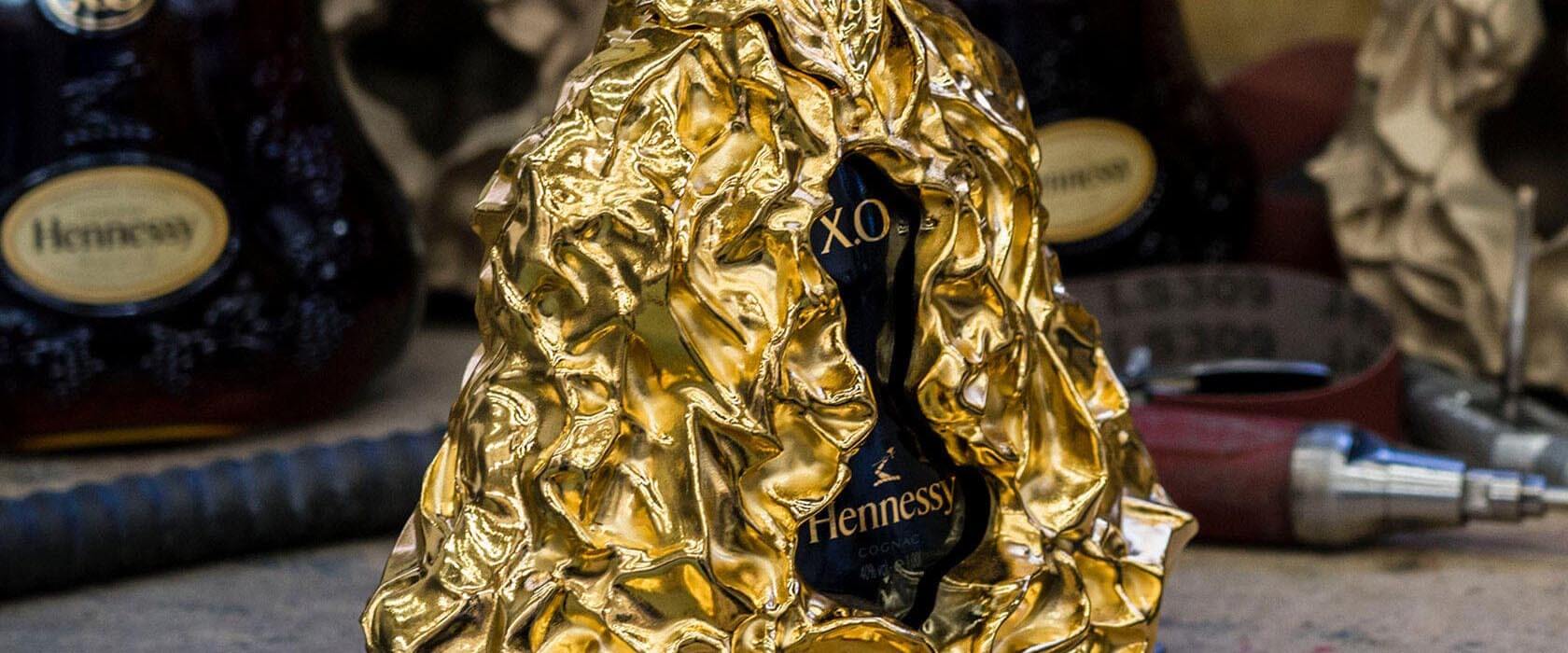 A Hennessy XO bottle partially encased in crumpled gold-colored metal. Other bottles and tools are blurred in the background. The design gives a luxury and artistic feel.