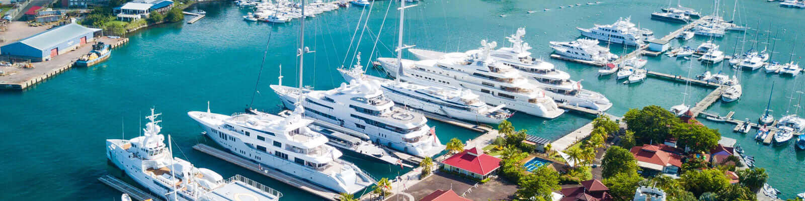 Arial view of a marina with several large white yachts docked beside a pier. Surrounding the marina are lush green trees, a few buildings with red roofs, and a clear blue sky reflecting on the water.