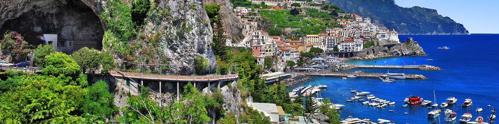 Scenic view of a coastal town with colorful buildings on a hillside overlooking the sea. A winding road passes through lush greenery and rocky cliffs. Boats are docked in the vibrant blue water, with a mountainous backdrop under a clear sky.