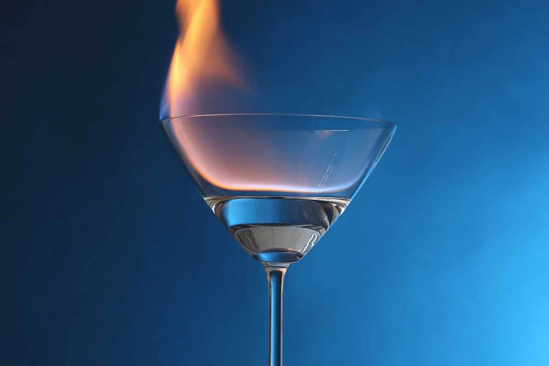 A martini glass filled with a clear liquid, topped with a flame, against a blue gradient background.