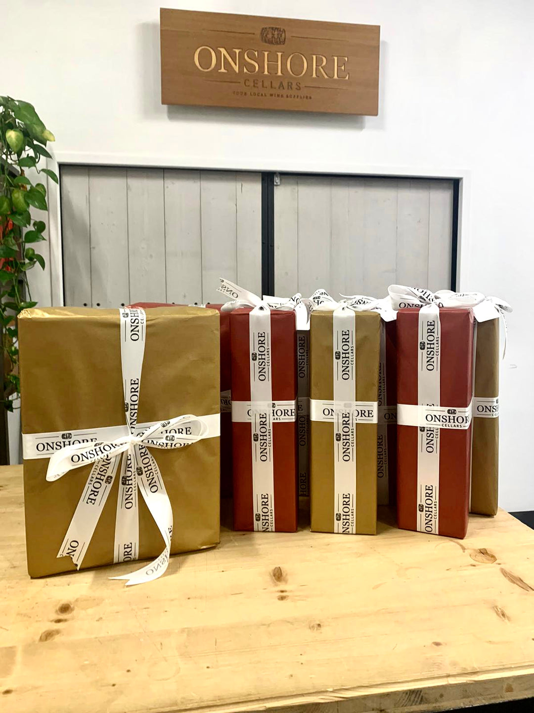 A display of gift-wrapped boxes in red and gold paper with white ribbon on a wooden table. A wooden sign in the background reads Onshore Cellars. A green plant is partially visible on the left.