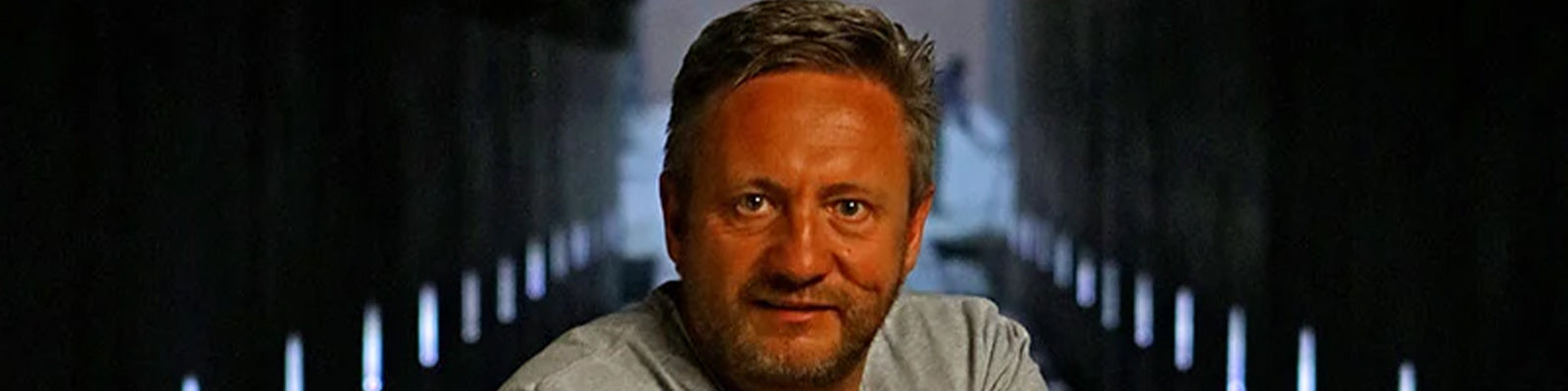 A person with short hair and a beard is sitting in a dimly lit, tunnel-like space with lights along the walls. He is wearing a gray shirt and looking at the camera.