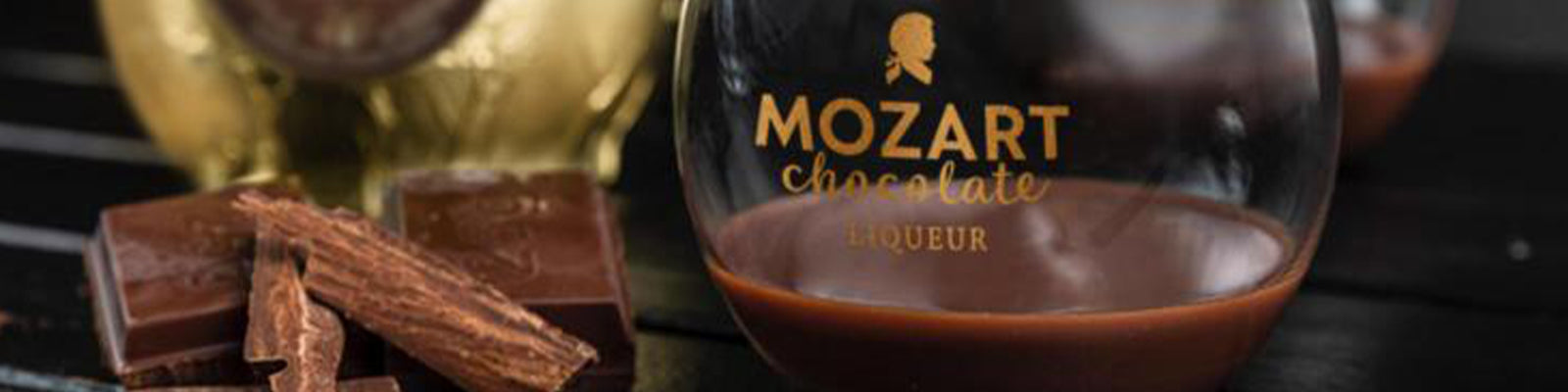 A glass of Mozart chocolate liqueur next to chocolate pieces on a dark surface.