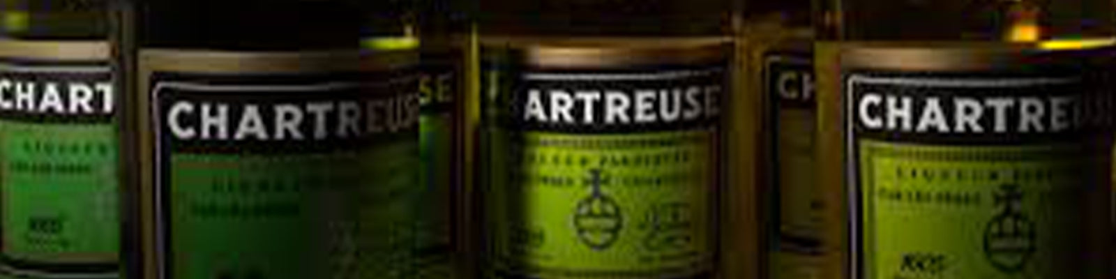 Close-up of several Chartreuse bottles with green labels. The labels have text and a circular emblem, illuminated under subtle lighting.