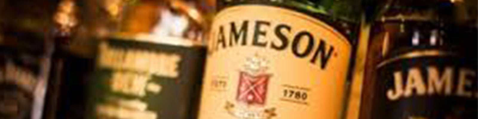 Close-up of several bottles of Jameson whiskey, with one label prominently displaying the name Jameson and a date of 1780. The background shows other blurred bottles, creating a warm, inviting atmosphere.