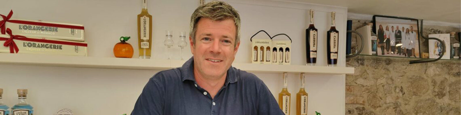 A person in a navy shirt stands smiling behind a counter, surrounded by bottles and packages on display shelves. The background features stone walls and framed photos.