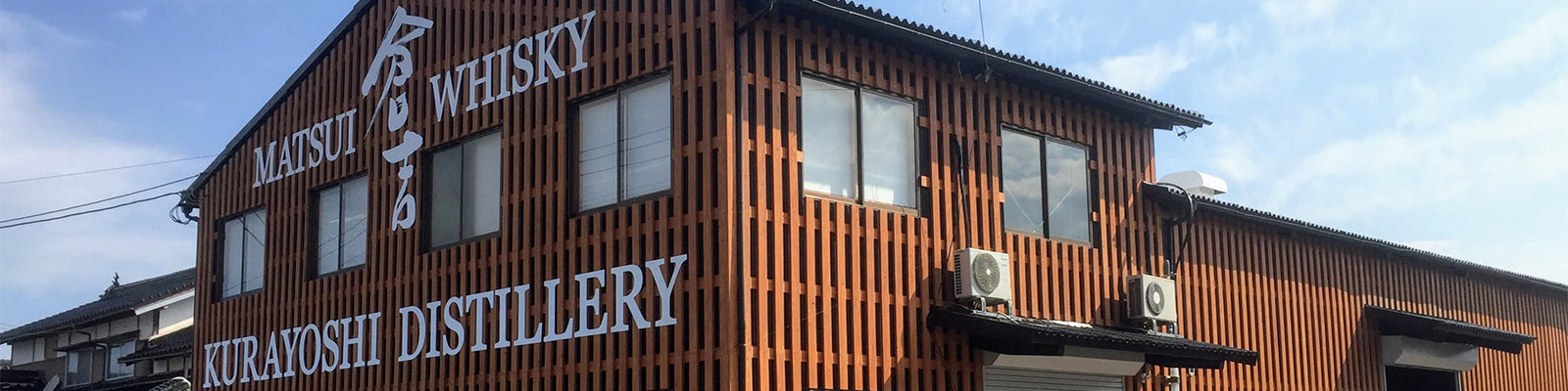 A wooden building with signage for Matsui Whisky Kurayoshi Distillery, featuring vertical slats and windows, against a blue sky.