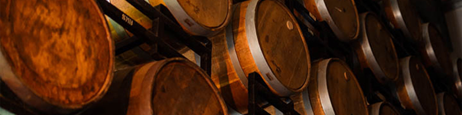 Rows of stacked wooden barrels used for aging wine or spirits in a dimly lit cellar. The barrels are arranged horizontally on shelves, displaying their rounded sides and metal bands.