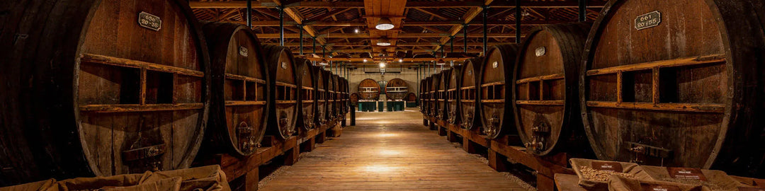 A dimly lit winery interior with large wooden barrels lined on both sides of a wooden walkway. The ceiling has exposed beams and lights, creating a rustic atmosphere.