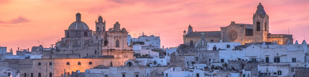 Vue pittoresque d'une ville historique avec des bâtiments blancs et des dômes ornés au coucher du soleil. Le ciel est rempli de teintes roses et orangées, créant une atmosphère chaleureuse et sereine.