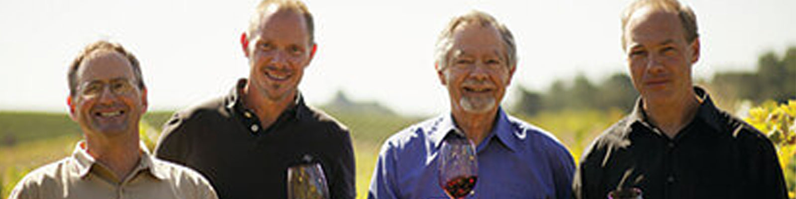 Four men standing outdoors in a vineyard, smiling and holding wine glasses. They are dressed casually, and the sunlit vineyard is in the background.